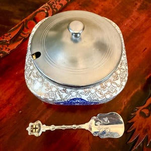 Op de afbeelding: Een vintage suikerpot met een zilveren deksel en een bijpassende lepel. De kom is wit met gouden bloempatronen en blauwe accenten. De lepel heeft een decoratief handvat met een wapen. De items staan op een donkerrode ondergrond.