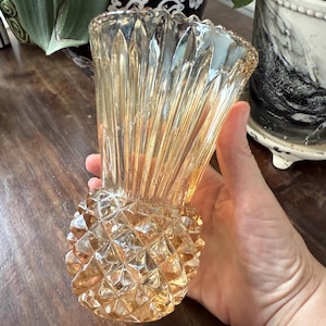 May include: An amber-coloured glass vase with a pineapple-shaped base and a fluted top. The vase is held in a hand, showcasing its diamond-cut pattern and warm, golden hue. The vase is approximately 15 cm tall.