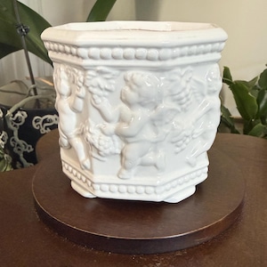 Peut inclure: Pot de fleurs en céramique blanche de forme hexagonale, orné de reliefs détaillés de chérubins. Le pot est posé sur un support en bois brun foncé, mettant en valeur le design et ajoutant de l'élégance.