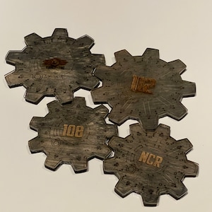 May include: Four wooden gears with laser-etched designs. The gears are gray with a wood grain pattern. The top gear has the text "Vault-Tec" in the center. The gear to the right has the text "112" in the center. The gear to the left has the text "108" in the center. The bottom gear has the text "NCR" in the center.