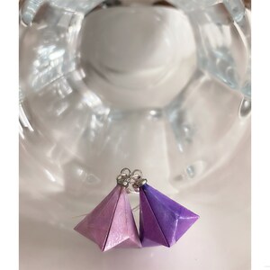 Peut inclure: Une paire de boucles d'oreilles en origami dans des tons de violet et de rose. Les boucles d'oreilles sont en papier et ont une forme géométrique. Elles sont attachées à des crochets d'oreille argentés.