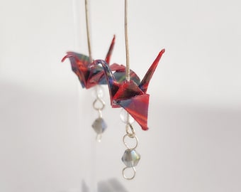 May include: Red and green origami crane earrings with a silver dangle charm. The earrings are made with paper and have a silver hook.