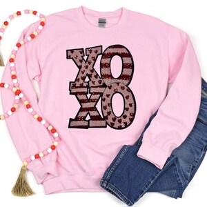 May include: Pink sweatshirt with a red and white sequined "XOXO" design. The sweatshirt has long sleeves and a crew neck.