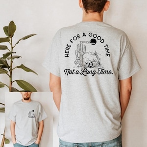 Puede incluir: Una camiseta gris con un estampado gráfico negro de un esqueleto en un paisaje desértico. El texto de la camiseta dice "Here for a good time, not a long time."