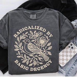 Puede incluir: Camiseta de manga larga gris oscuro con el texto "RADICALIZED BY BASIC DECENCY" en un diseño circular con un pájaro y elementos florales. Se ve un teléfono con una funda a cuadros.