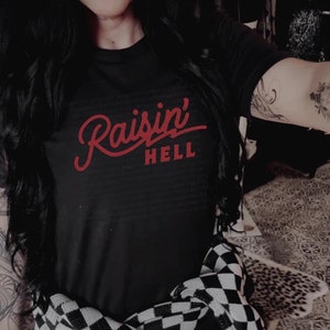 Puede incluir: Una camiseta negra con el texto rojo "raisin' hell" en la parte delantera. La camiseta la lleva puesta una persona con el pelo largo y negro y una bufanda a cuadros.