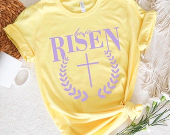 Könnte beinhalten: Gelbes T-Shirt mit dem Text "He is Risen" in lilafarbener Schrift mit einem lilafarbenen Kreuz- und Kranzdesign.
