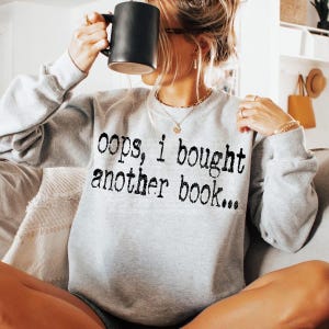 Pode incluir: Um moletom cinza claro com o texto "oops, I bought another book..." em preto. Uma caneca preta é segurada em frente ao rosto. A pessoa está usando joias de ouro.