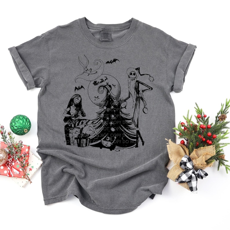 May include: A dark gray t-shirt with a black and white graphic print of Jack Skellington and Sally from The Nightmare Before Christmas standing in front of a Christmas tree. The graphic also includes a moon and bats.