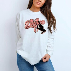 Op de afbeelding: Witte longsleeve trui met een roze strik en het woord "Dance" in een gestileerd lettertype. Een silhouet van een danseres bevindt zich in de strik.
