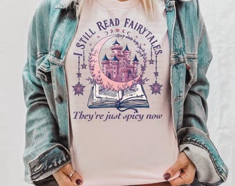May include: A pink t-shirt featuring a graphic of a castle in a crescent moon with stars and a book. The text reads "I still read fairytales, they're just spicy now".
