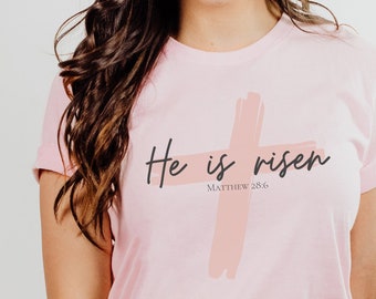 May include: A pink t-shirt with a pink brushstroke cross and the text "He is risen" with the scripture reference "Matthew 28:6".