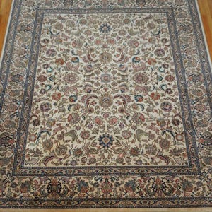 May include: A rectangular area rug featuring an intricate floral and vine pattern in shades of cream, blue, red, and green. The design includes a central medallion motif surrounded by a detailed border and finished with short fringe on the ends.