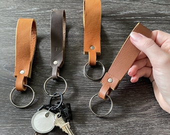 May include: Four leather keychains with metal rings. Two are brown, one is dark brown, and one is a lighter brown. The keychains are on a dark wood surface. Two keys are attached to one of the keychains.