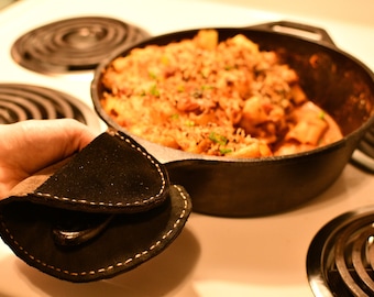 May include: A person is holding a black leather pot holder with a white stitch detail. The pot holder is being used to lift a cast iron skillet with a handle. The skillet is filled with a creamy, orange-colored dish.