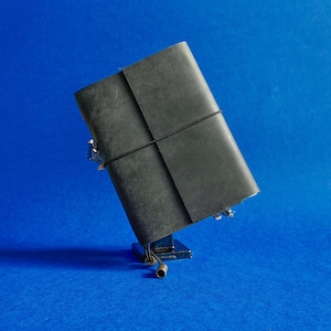 May include: A black leather journal with a black elastic closure. The journal is held in place by a small, silver-colored metal stand. The background is a solid blue.