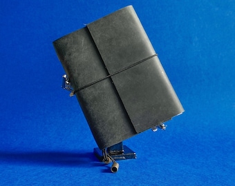 May include: A black leather journal with a black elastic closure. The journal is held in place by a small, silver-colored metal stand. The background is a solid blue.