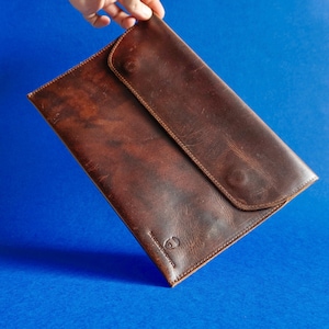 May include: A brown leather document holder with a flap closure. The leather has a distressed appearance with variations in color. The document holder is rectangular with rounded corners and a stitched border. The background is a solid blue.