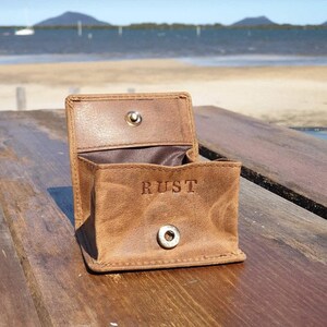 May include: Open, brown leather coin purse with the word "RUST" embossed on the front. The purse is open, revealing a brown interior. It has a snap closure and is sitting on a wooden surface.