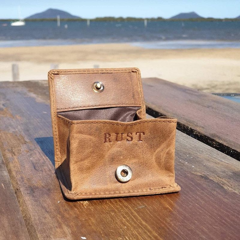 May include: Open, brown leather coin purse with the word "RUST" embossed on the front. The purse is open, revealing a brown interior. It has a snap closure and is sitting on a wooden surface.