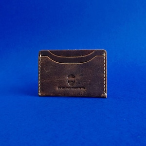 Może przedstawiać: Brązowy skórzany etui na karty z białymi przeszyciami i logo. Etui na karty ma dwie przegródki na karty i kieszeń. Tekst "THE LEATHER TRADING CO." jest wydrukowany na dole.