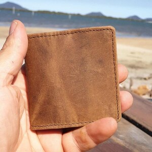 May include: A square, brown leather wallet with visible stitching around the edges. The wallet is held in a hand, with a beach and ocean in the background. The leather has a distressed appearance.