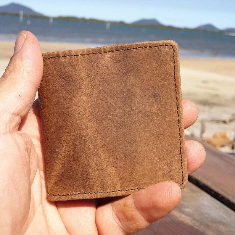 May include: A square, brown leather wallet with visible stitching around the edges. The wallet is held in a hand, with a beach and ocean in the background. The leather has a distressed appearance.