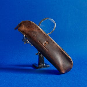 Könnte beinhalten: Ein braunes Lederetui mit Reißverschluss, das eine Lupe mit silbernem Rahmen enthält. Das Etui hat ein Logo und Text aufgedruckt. Der Hintergrund ist einfarbig blau.