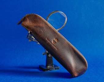 May include: A brown leather case with a zipper, holding a magnifying glass with a silver frame. The case has a logo and text printed on it. The background is a solid blue.