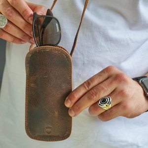 May include: A brown leather sunglasses case with a neck strap, holding a pair of sunglasses. The case has a distressed finish and a small logo at the bottom. The sunglasses have dark frames and lenses.