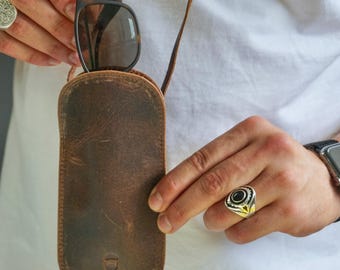 May include: A brown leather sunglasses case with a neck strap, holding a pair of sunglasses. The case has a distressed finish and a small logo at the bottom. The sunglasses have dark frames and lenses.
