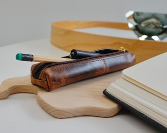 May include: A brown leather pencil case with a gold zipper, holding a pencil and a black pen, rests on a wooden cutting board. A notebook and a wooden tray are in the background. The pencil has a green eraser.
