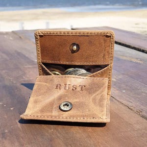May include: Open brown leather coin purse with the word "RUST" embossed on the front flap. The purse is open, revealing coins inside. The purse has a snap closure and is sitting on a wooden surface.