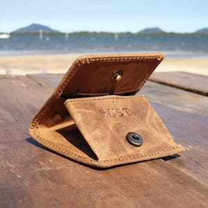 May include: Open, brown leather wallet with the word "RUST" embossed on the inside flap. The wallet is open, revealing the interior. A metal snap closure is visible. The wallet is on a wooden surface.