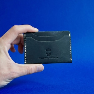 Może przedstawiać: Czarny skórzany etui na karty z białymi przeszyciami i wieloma przegródkami na karty. Etui na karty ma napis "THE LEATHER TRADING CO." i logo. Etui na karty jest trzymane na niebieskim tle.
