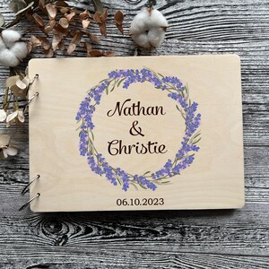 Könnte beinhalten: Ein Gästebuch aus Holz mit einem Design aus Lavendelkränzen. Das Buch ist personalisiert mit den Namen "Nathan & Christie" und dem Datum "06.10.2023".