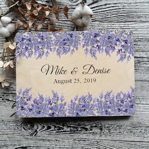 Può includere: Un libro degli ospiti in legno con un design floreale di lavanda. Il libro ha una copertina in legno chiaro con il testo "Mike & Denise August 25, 2019" stampato su di essa.