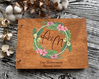 Könnte beinhalten: Ein Holzfotoalbum mit einem floralen Kranzdesign und den Initialen "A & M" in einer kursiven Schrift. Der Text "Our Honeymoon Hawaii 2024" ist unter dem Kranz gedruckt.