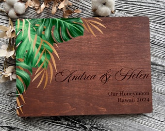 Könnte beinhalten: Ein braunes Holz-Gästebuch mit einem tropischen Blattdesign und dem Text "Andrea & Helen Our Honeymoon Hawaii 2024" auf der Vorderseite.