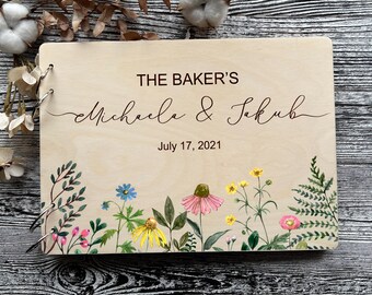 Könnte beinhalten: Ein Gästebuch aus Holz mit Metallbindung. Der Einband ist aus hellem Holz, graviert mit "THE BAKER'S" über den Namen "Michaela & Jakub" und dem Datum "July 17, 2021". Der untere Rand zeigt ein Aquarell-Blumenmuster.