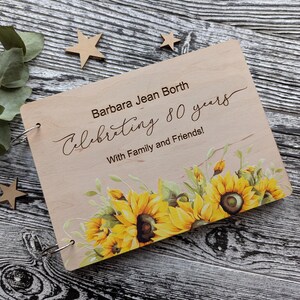 May include: A wooden photo album with a sunflower design. The cover is engraved with the text "Barbara Jean Borth Celebrating 80 years With Family and Friends!"