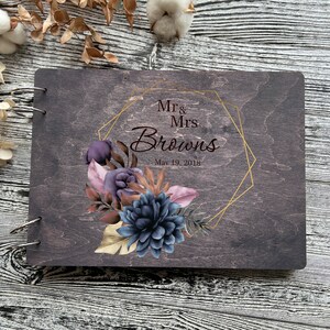 May include: A dark wood guest book with a gold geometric frame and floral design. The text reads "Mr. & Mrs. Browns May 19, 2018".