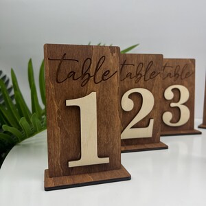 May include: Wooden table number signs with the word "table" and a number on each sign. The numbers are 1, 2, and 3. The signs are brown with a light brown wood grain.