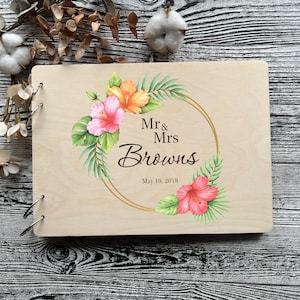 Puede incluir: Un libro de visitas de madera con un diseño floral con flores de hibisco rosa y naranja, hojas verdes y un círculo dorado con el texto "Mr & Mrs Browns May 19, 2018".