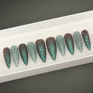 May include: Ten green and brown long, pointed false nails with a textured, iridescent finish. The nails are arranged in a row on a white surface.