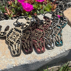 May include: Five wooden keychains shaped like a bull skull with geometric patterns. The keychains are in various colors, including brown, white, teal, and pink.