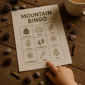 Puede incluir: Una tarjeta de juego "Mountain Bingo" con ilustraciones de elementos de la naturaleza como piñas y hojas. El dedo de un niño señala la casilla "Twig". La escena incluye una taza de café, pequeñas piedras y un árbol de fieltro sobre una superficie de madera.