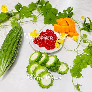 May include: A still-life image featuring a green bitter melon, sliced pieces, red seeds, and orange pieces. Yellow flowers and green vines are arranged around the food. The text "Madflower Trail" and "Bitter Melon" are visible.