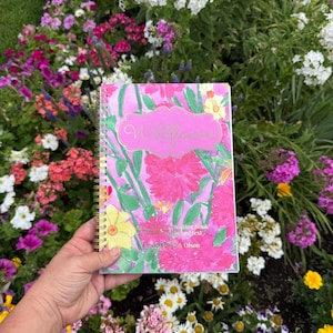 May include: A spiral-bound notebook with a vibrant pink and green floral watercolor cover. The front features a decorative label with the text Wildflowers, followed by Watercolor Painting Book with drawings and text by Nancy Audack Olson.