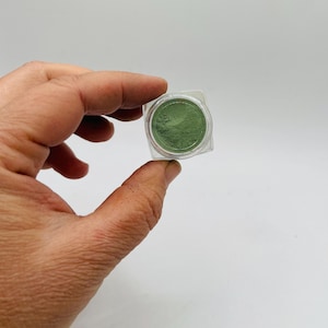 May include: A small, clear plastic container holding a vibrant green powder. The container is square with rounded corners. The powder appears finely milled and evenly distributed within the container. The background is a plain white surface.